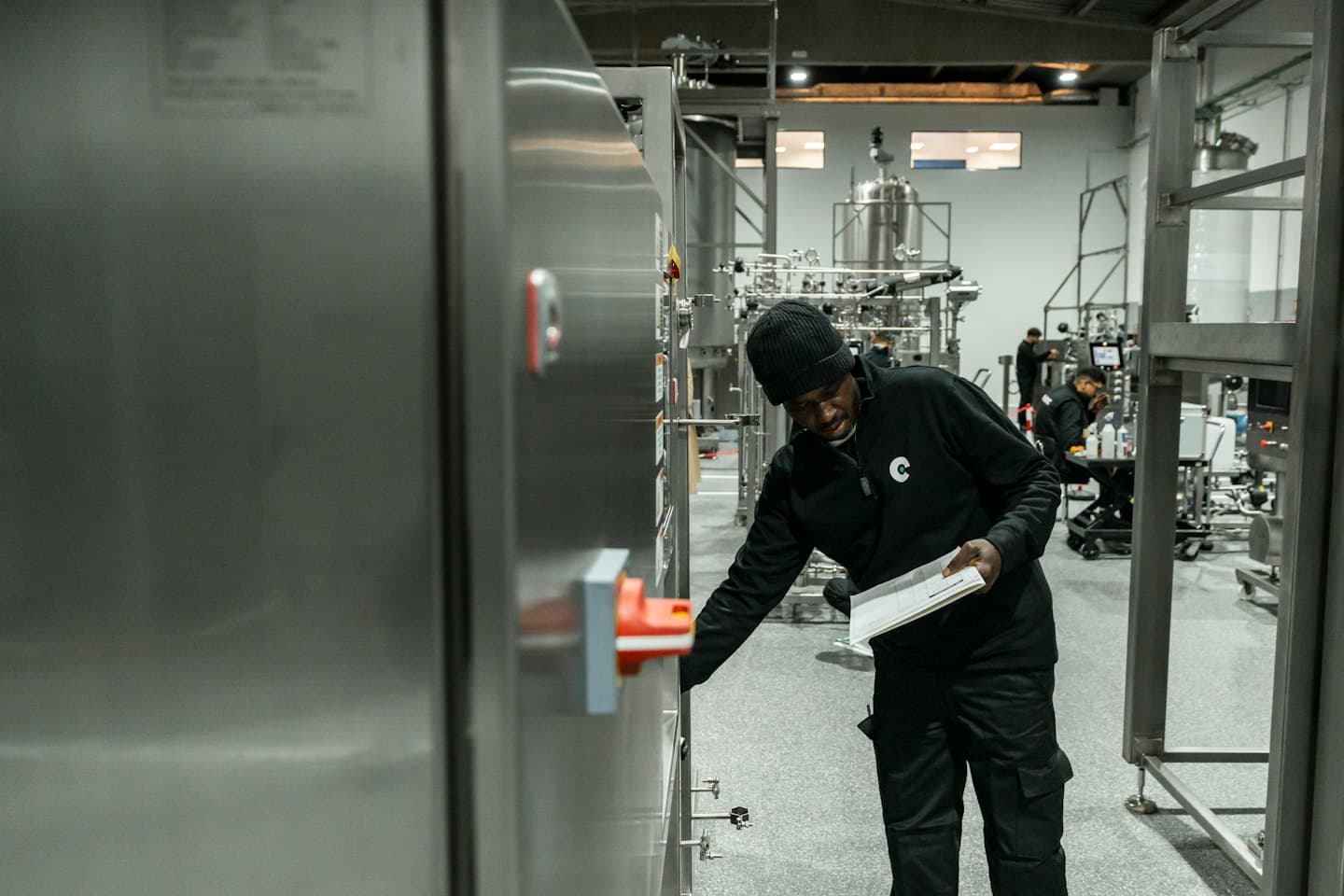 Worker in uniform inspects industrial control panel with clipboard.